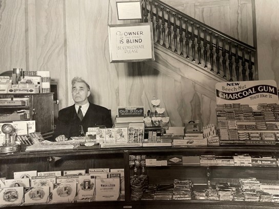 a black-and-white photo of a man sitting behind a store counter, with a sign that says "the owner of this stand is blind, be considerate please."