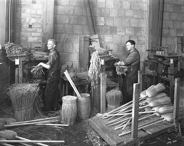 A black-and-white photo of two men at workstations, making brooms.