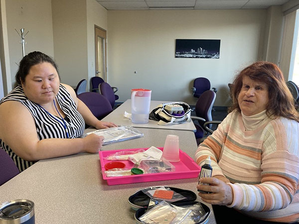 Kia and Charlene, two SSB staffers, sit at a table across from each other with Low Vision training tools.