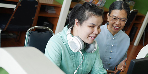 person with headphone sitting at computer with assistance from another person